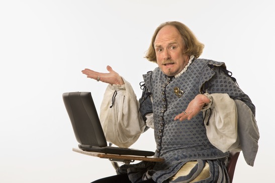 William Shakespeare in period clothing sitting in school desk with laptop computer shrugging at viewer.