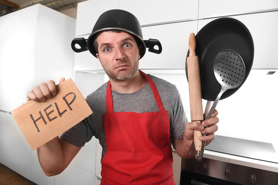 funny 30s Caucasian man holding pan and household with pot on his head in red apron at home kitchen asking for help unable to cook showing panic on cooking with funny face expression