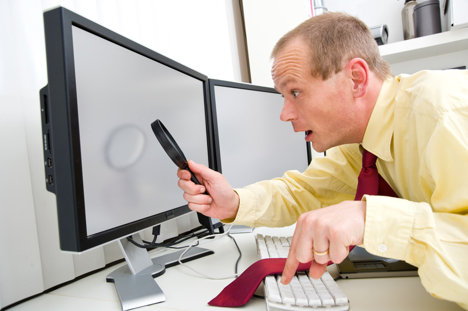 A man being surprized while scruitining  a computer monitor with a magnifying glass, his left index finger hovering above the Escape key