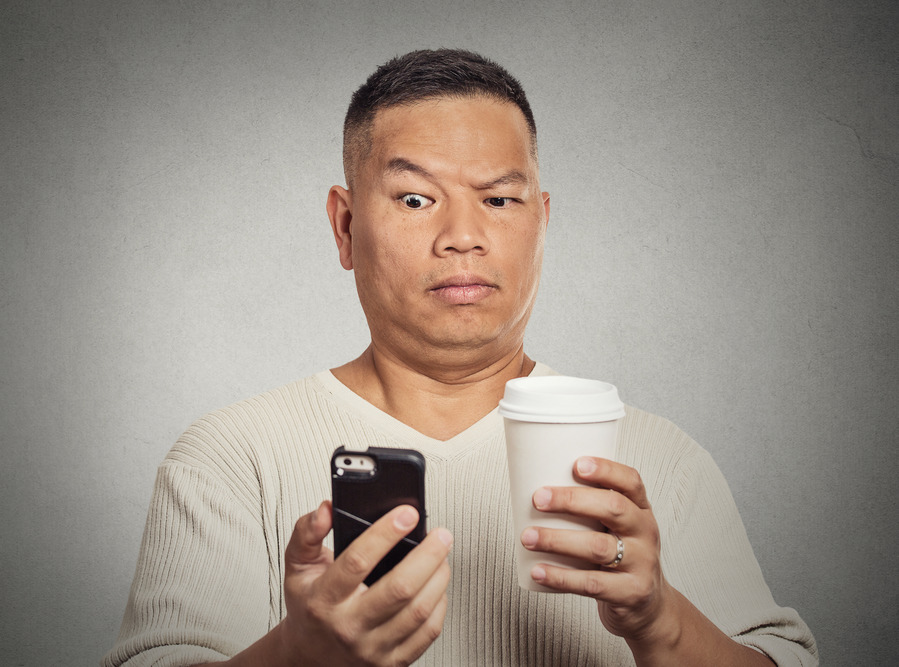 Closeup portrait worried skeptical surprised man reading bad news sms on smart mobile phone drinking holding cup coffee isolated grey wall background. Human face expression emotion  reaction