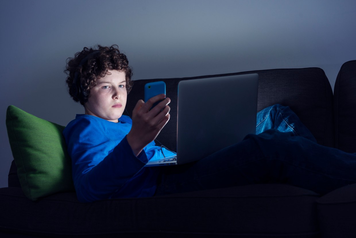 Portrait of a young teenage boy sitting on the sofa at home texting on a smartphone and watching something on screen at the same time. He is illuminated by the light from the screen whilst everything else around him is dark.