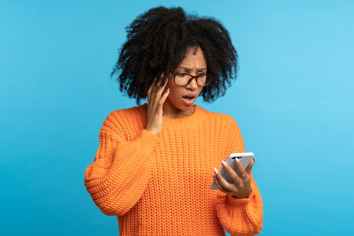 Studio portrait of confused shocked African ethnicity woman looking at smartphone with opened mouth, reading bad news, notification, unpleasant message isolated on blue color background.