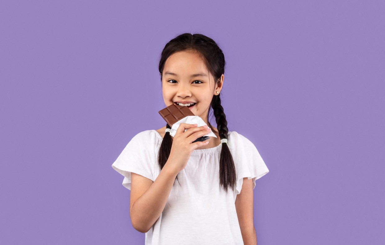 Sweet Tooth. Little Asian Girl Eating Chocolate Smiling To Camera Standing On Purple Background. Studio Shot