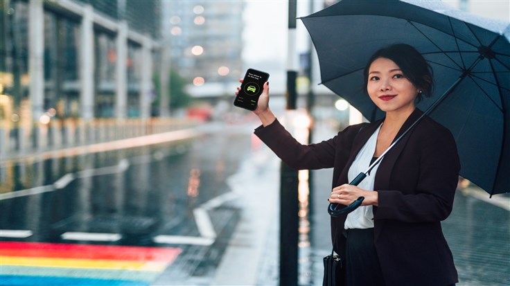 Young Asian businesswoman arranging taxi service using mobile app on smart phone, holding an umbrella in a rainy day in the city. Business on the go concept.