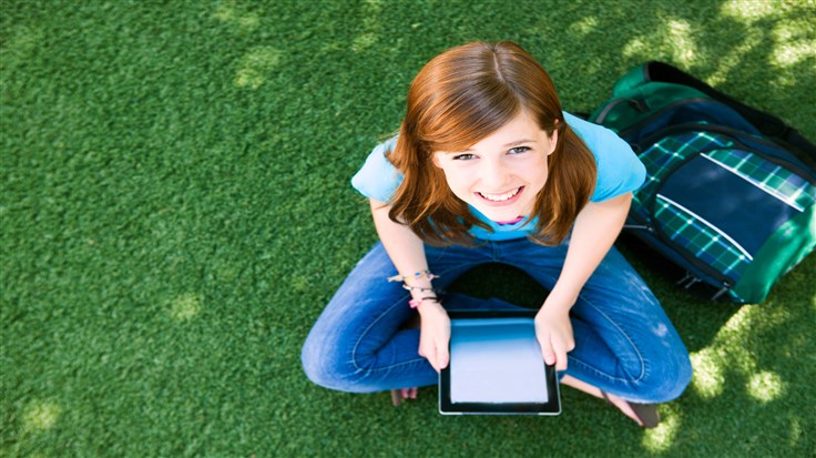 Overhead shot of a beautiful young red-headed girl on the grass with a backpack and a digital tablet