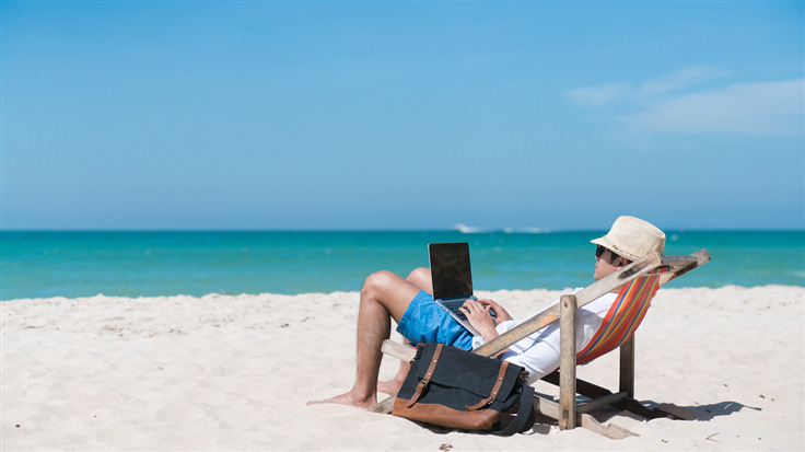 A man in shorts and a hat looks at his laptop while sitting on the beach, the ocean before him