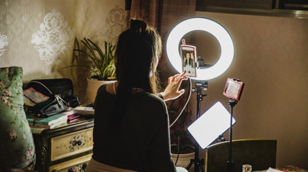 A woman live-streams herself using her smart phone and a ring light. The woman is shown from behind.