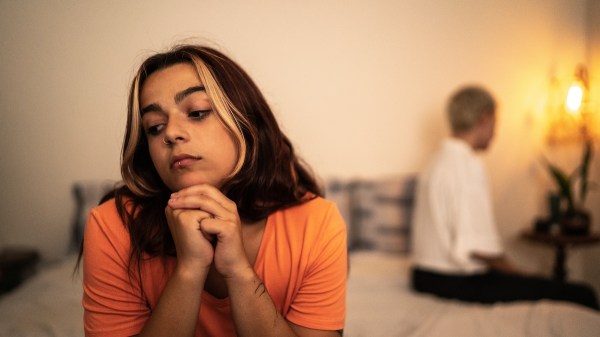 A couple sits apart on a bed, facing different directions. A woman, closer to the camera, looks pensive.