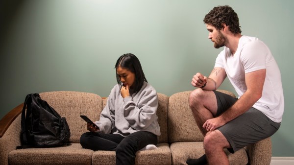 A man sits atop a couch's armrest as his romantic partner sits looking at her phone. The two are not facing one another and seem tense.