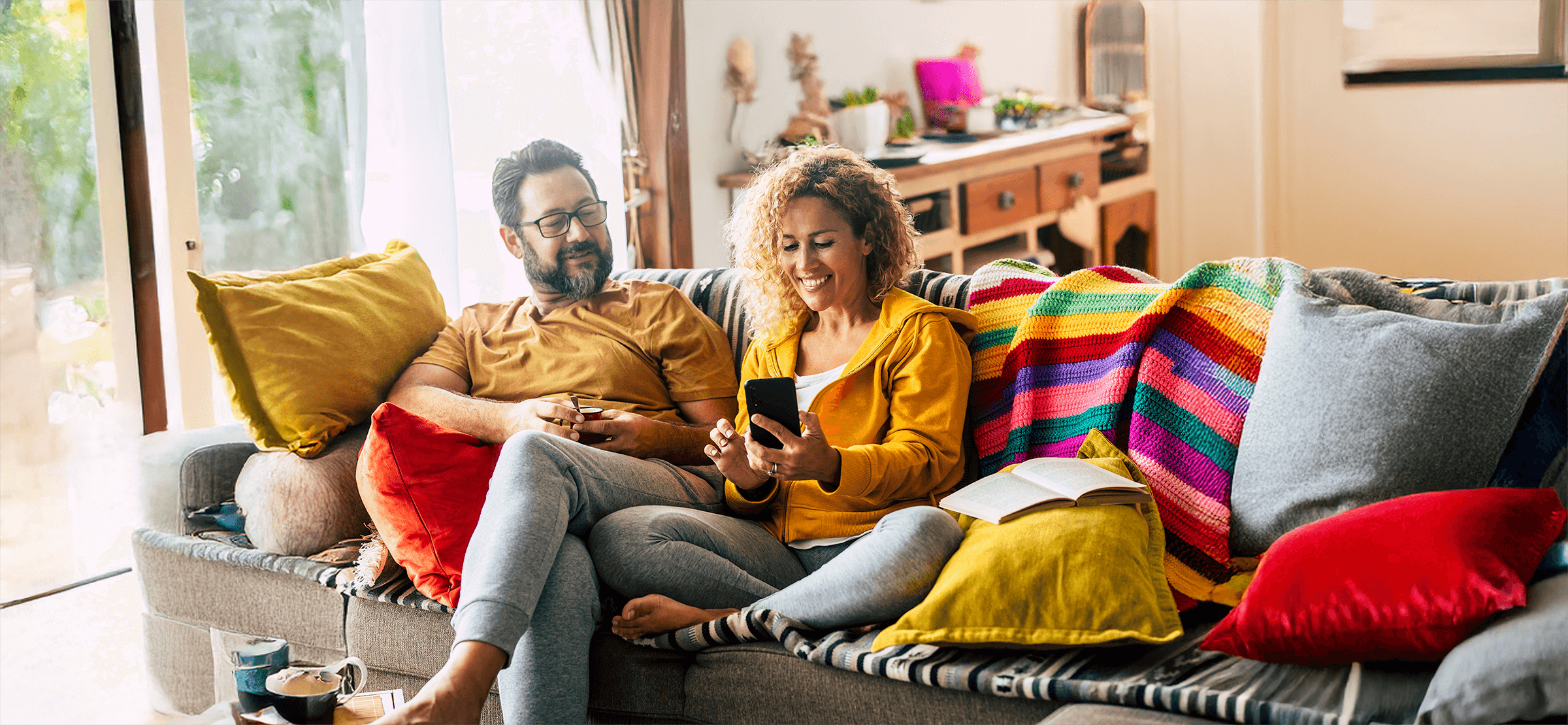 Zwei Menschen sitzen in einem farbenfrohen Wohnzimmer, vergleichen ihre Mobilgeräte und lächeln
