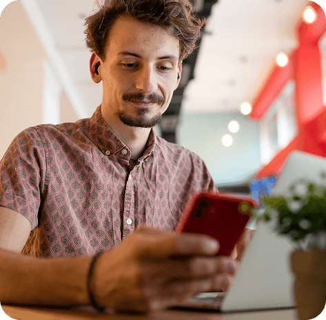Young masculine person sitting in a cafe with a laptop and looking at a red smartphone